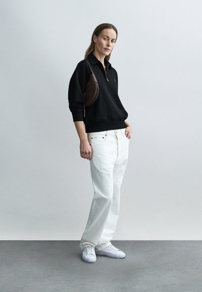 Svart genser med halvglidelås, hvite vide jeans, hvite joggesko og en liten brun veske. Glatte teksturer og et monokromatisk fargevalg.