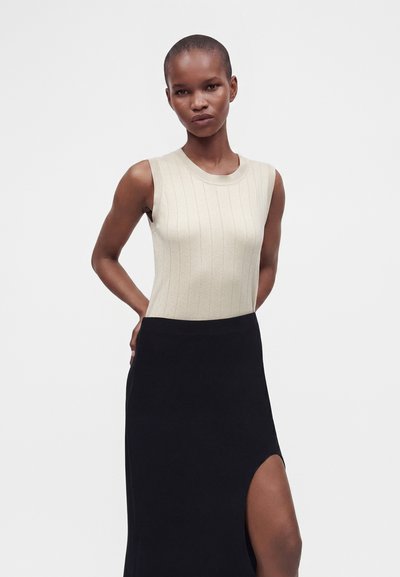 Woman with short hair wearing a sleeveless beige ribbed top and a black skirt with a thigh-high slit, standing against a plain background.