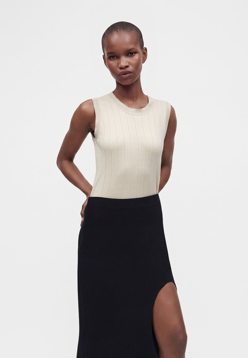 Femme aux cheveux courts portant un haut côtelé beige sans manches et une jupe noire avec une fente haute sur la cuisse, debout devant un fond uni.