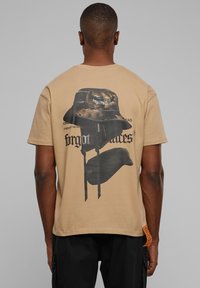 Beige t-shirt med et grafisk print af en camouflage bøttehat og sort tekst på ryggen, med en lige pasform og et sideetiket.