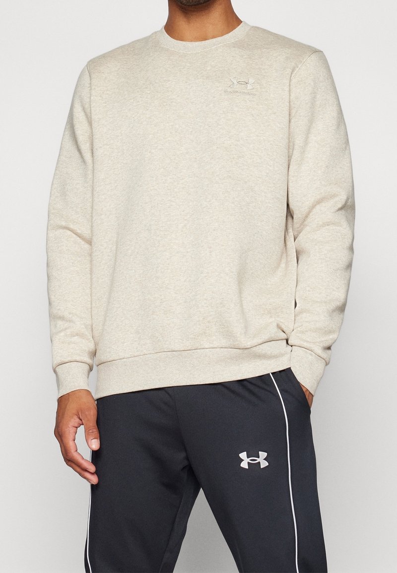 Man som bär en beige sweatshirt med rund hals och en liten logotyp samt svarta sportbyxor med kontrasterande sidolinjer och logotyp.