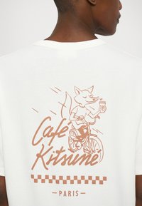 Biała bawełniana koszulka z brązowym nadrukiem przedstawiającym lisa jadącego na rowerze, trzymającego napój, z napisem "Café Kitsuné Paryż" poniżej.