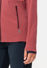 Giacca in pile color marrone con tessuto testurizzato, tasca sul petto con zip e polsini elastici. Abbinata a pantaloni blu scuro con dettagli a pannello.