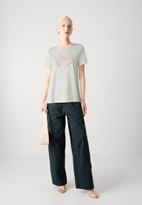 T-shirt vert clair avec un motif de nœud rose, associé à un pantalon sombre ample. Le mannequin tient un sac beige clair.