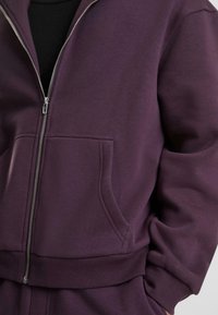 Lilla zip-up hettegenser med en glatt tekstur, sidelommer og ribbede mansjetter. Har et lite designelement under glidelåsen.
