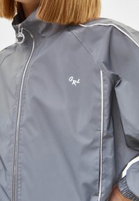 Šedá windbreaker bunda s zapínáním na zip, bílými lemovacími detaily a malým logem "GRL" na hrudi. Hladký, lehký materiál.