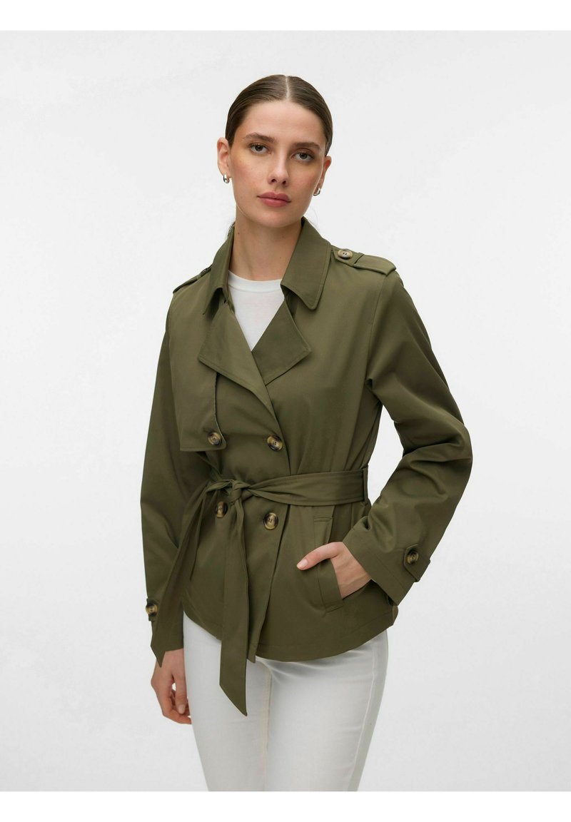 Femme portant une veste croisée ceinturée olive avec des boutons, une chemise blanche en dessous et un pantalon blanc sur un fond uni.