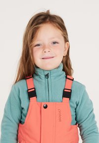 Peto de color coral con tirantes negros ajustables sobre una chaqueta de forro polar verde azulado. La chaqueta tiene un cuello alto y un cierre de cremallera.