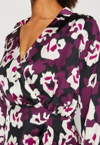 Gros plan sur une robe portefeuille à motif floral violet, noir et blanc, avec un col en V et une taille froncée portée par une personne.