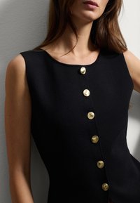 Schwarzes ärmelloses Oberteil mit Rundhalsausschnitt, ausgestattet mit glänzenden goldenen Knöpfen auf der Vorderseite und einem strukturierten Stoff. Minimalistisches Design.