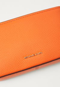 Orange läderplånbok med texturerad yta, prydd med guld "Michael Kors"-logotyp och kontrasterande kantstickning. Slät inredning.