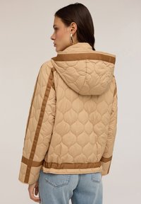 Giacca trapuntata beige con cappuccio, caratterizzata da accenti marroni e un design squadrato. La texture è morbida e l'orlo presenta una fascia marrone solida.