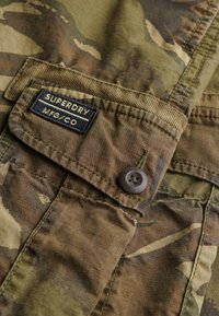 Kamuflage stof skjorte med en brun knap, der har et broderet "SUPERDRY MFG/CO" mærke på brystlommen. Struktureret materiale.