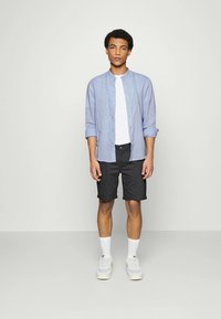 Camisa de botón azul claro sobre una camiseta blanca, combinada con pantalones cortos oscuros y zapatillas blancas, de pie sobre un fondo neutro.
