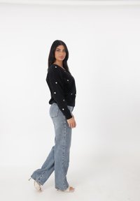 Schwarzer Pullover mit goldenen Herzmustern, kombiniert mit hellblauen, weit geschnittenen Jeans und weißen High Heels. Klarer Hintergrund.