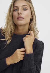 Donkergrijs gebreid sweater met ribbeltextuur, ronde halslijn en donzige manchetten. Model houdt de sweater voorzichtig bij haar schouder.