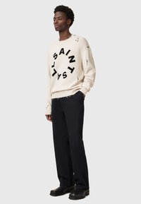 Off-White Strickpullover mit schwarzer "SAINT" Schrift und ausgewaschenen Details, kombiniert mit schwarzen Hosen und schwarzem Schuhwerk.
