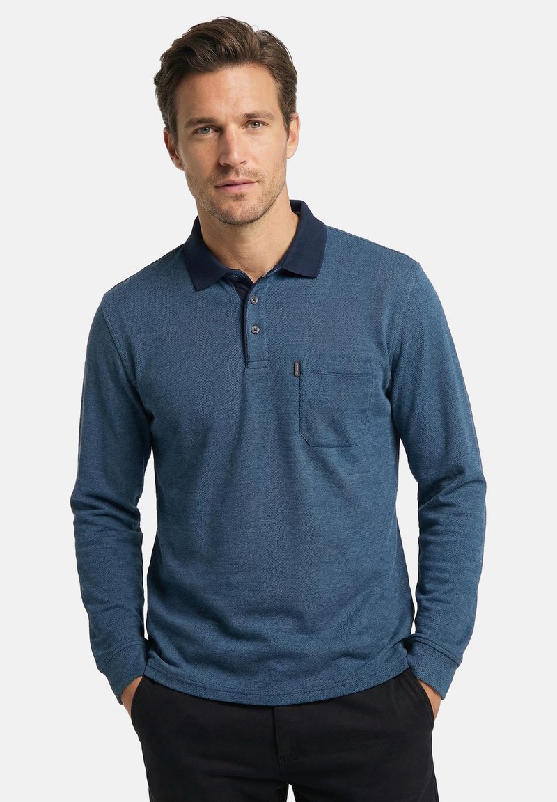 Mann trägt ein blaues, langärmliges Poloshirt mit Brusttasche, marineblauem Kragen und schwarzer Hose, steht mit den Händen in den Taschen.