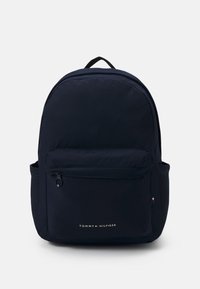 Zaino in nylon blu marino con parte superiore curva, tasca frontale con zip e tasca laterale. Presenta un manico superiore e il logo Tommy Hilfiger in bianco.