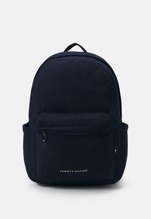 Zaino in nylon blu marino con parte superiore curva, tasca frontale con zip e tasca laterale. Presenta un manico superiore e il logo Tommy Hilfiger in bianco.
