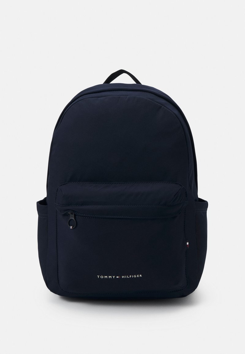 Zaino in nylon blu marino con parte superiore curva, tasca frontale con zip e tasca laterale. Presenta un manico superiore e il logo Tommy Hilfiger in bianco.