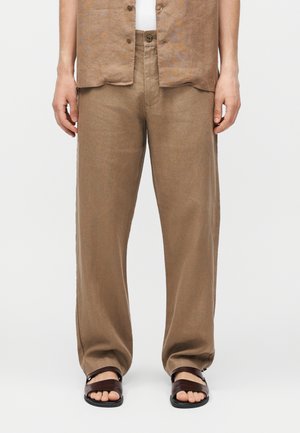 Personne portant un pantalon marron ample, des sandales marron, une chemise à manches courtes marron clair et un sous-vêtement blanc, debout devant un fond uni.
