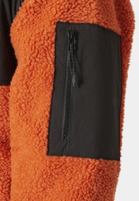 Fleece bunda s oranžovým texturovaným povrchem a černou látkovou panelem se zipovou kapsou na rukávu.