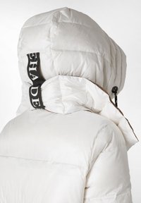 Hvid pufferjakke med hætte. Har en struktureret overflade, vandret syninger, kontrasterende brun kant og et sort logo bånd på kraven.