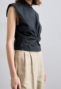 Camiseta negra sin mangas con cuello tipo polo, que presenta un diseño frontal retorcido, cierres de botones y una textura de tela suave. Se lleva con pantalones plisados beige.