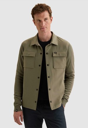 Homme aux cheveux courts et bouclés portant une veste olive boutonnée avec des poches à rabat, une chemise noire et un pantalon foncé, sur un fond uni.