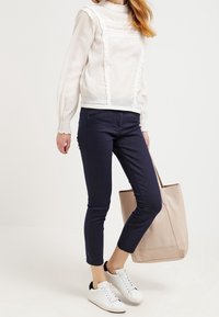 Blusa blanca de manga larga con detalles de volantes, pantalones oscuros de corte slim en azul marino y zapatillas blancas con acentos negros. Se sostiene un bolso de tote beige.