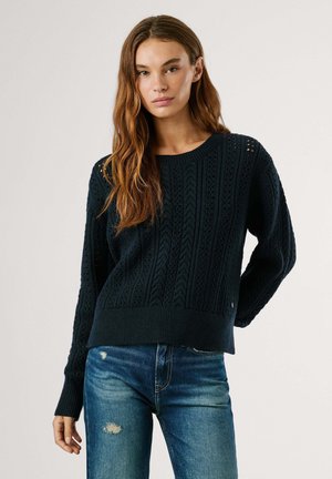 Diepblauwe gebreide trui met een kabelpatroon en geribde manchetten, gecombineerd met versleten blauwe jeans. De trui heeft een losse pasvorm.
