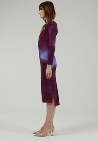 Vestido largo y ajustado con un patrón degradado en tonos morados y azules, con mangas largas y aberturas laterales para permitir el movimiento. Sandalias transparentes con tacón.