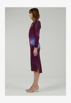Long, fitted dress with a gradient pattern in purple and blue shades, featuring long sleeves and side slits for movement. Transparent heeled sandals.