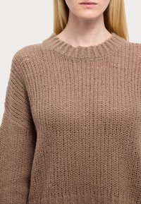 Femme portant un pull en maille épaisse marron avec un col rond côtelé et des manches longues, visible du cou jusqu'au milieu du torse.