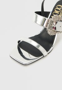 Ασημένιο πέδιλο με ψηλό τακούνι και τετράγωνη μύτη, δύο λουράκια και διακοσμητική αγκράφα με το λογότυπο Versace Jeans Couture.