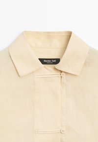 Beżowy kołnierzyk koszuli z ukrytą plisą na guziki i dwoma widocznymi beżowymi guzikami, z czarną metką Massimo Dutti wewnątrz kołnierza.