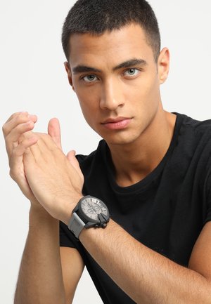 Black analog watch with a large round face, gray silicone band, and silver accents. Worn on a wrist with hands clasped together.