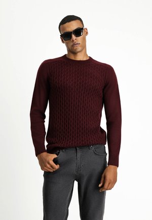 Maglione a maglia bordeaux con motivo a onde testurizzate, scollo rotondo e polsini a coste, abbinato a jeans grigi scuri. Occhiali da sole indossati.
