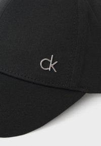 Svart cap med buet brem, laget av strukturert stoff, med en sølvfarget metall "ck" logo på venstre frontside.
