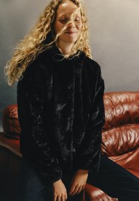 Czarna kurtka z pluszowego futra syntetycznego z kapturem, o luźnym kroju i miękkiej teksturze. Nosi ją osoba w ciemnych dżinsach, na stonowanym tle.