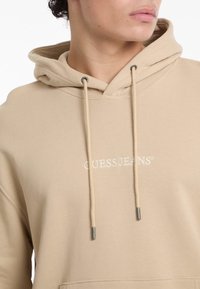 Felpa beige in cotone con tasca frontale, collo con cordino e logo ricamato "GUESS JEANS". Tessuto strutturato.