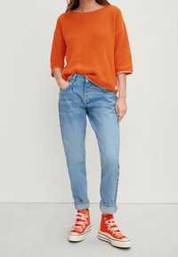 Suéter de punto texturizado naranja brillante con mangas anchas, combinado con jeans de tiro alto azul claro y zapatillas altas rojas.