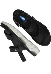 Sandales noires avec une tige en daim souple, des sangles réglables et une semelle en caoutchouc blanc texturé. Elles ont un bout arrondi et un amorti au niveau de la semelle.