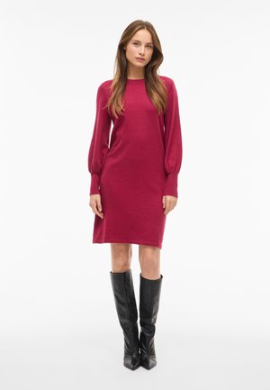 Femme debout portant une robe framboise à manches longues, longueur genou, avec des manches bouffantes et des bottes en cuir noires montant jusqu'au genou.