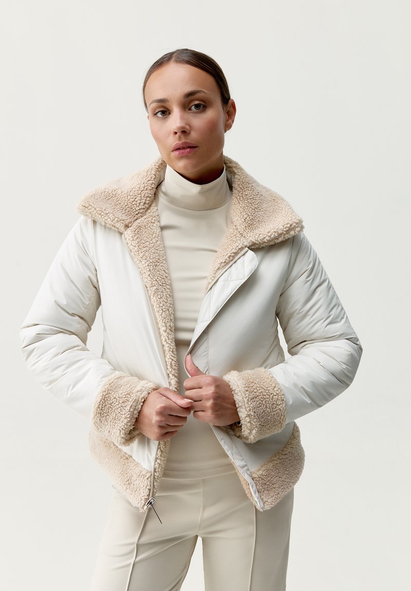 Biała kurtka puchowa z beżowym futrzanym kołnierzem i mankietami, z suwakiem i luźnym krojem, noszona na beżowym golfie.