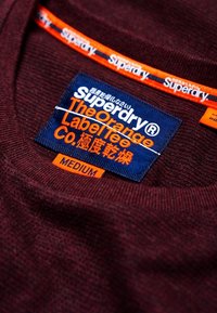Burgundy neulepuseron kaulus, jossa on sininen etiketti valkoisella ja oranssilla tekstillä, jossa lukee "Superdry", "The Orange Label Tee" ja "Medium."