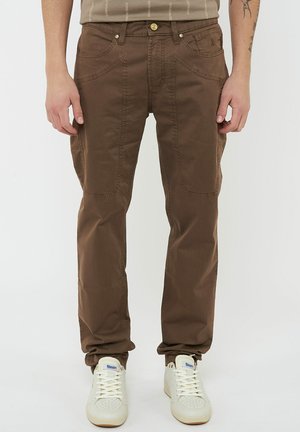 Uomo che indossa pantaloni marroni a gamba dritta con tasche frontali applicate e scarpe da ginnastica bianche, in piedi davanti a uno sfondo semplice.