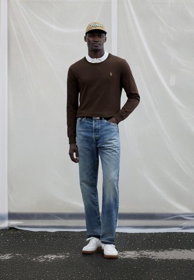 Brown long-sleeve sweater, white collared shirt beneath, light blue denim jeans, white sneakers with tan soles, brown cap with logo.