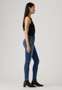 Νεαρή γυναίκα σε προφίλ, φορώντας μαύρο αμάνικο τοπ, σκισμένο στενό μπλε τζιν και μαύρα μποτάκια μέχρι τον αστράγαλο, μπροστά σε απλό φόντο.